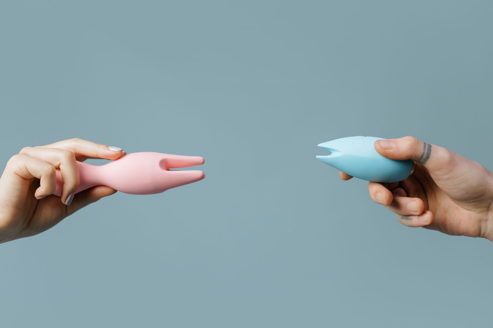 Two hands holding pink and blue silicone vibrators against a pastel background, representing reclaiming pleasure together